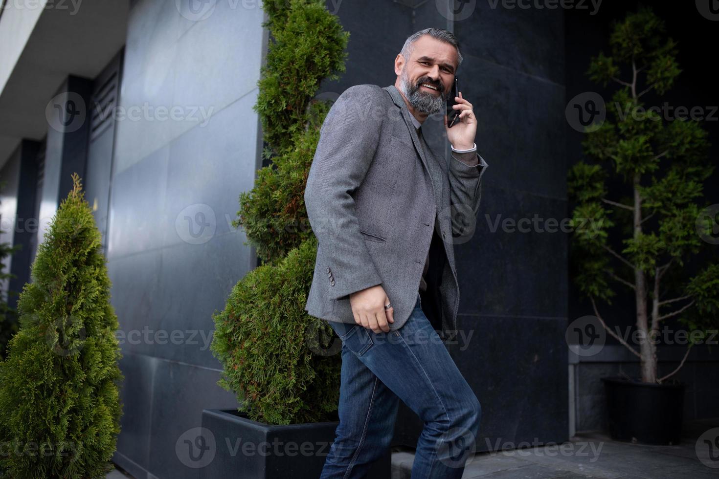 um empresário idoso entra em um prédio de escritórios falando em um telefone celular olhando ao redor com um sorriso foto