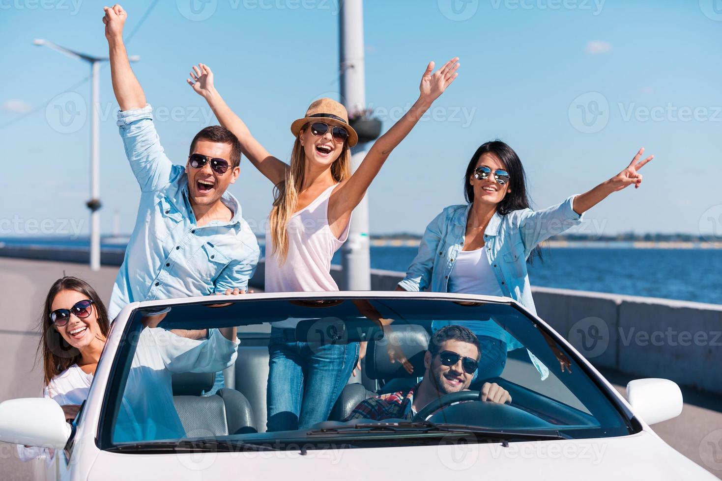 passando muito tempo no conversível. grupo de jovens felizes curtindo a viagem em seu conversível branco e levantando os braços foto