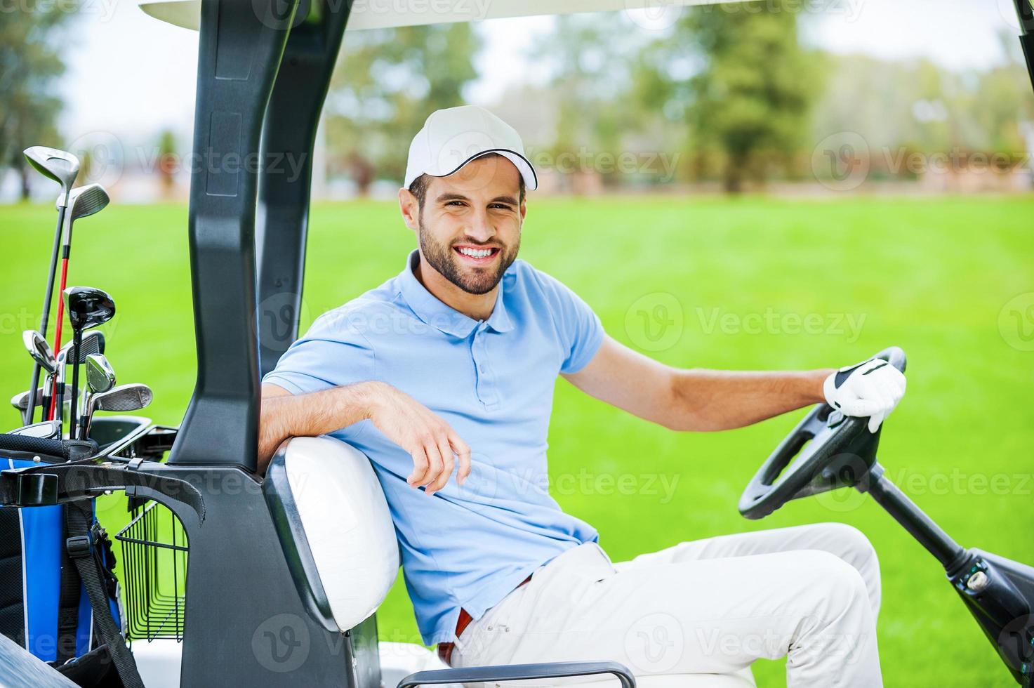 no meu caminho para o próximo buraco. Vista lateral do jovem jogador de golfe masculino feliz dirigindo um carrinho de golfe e olhando para a câmera foto