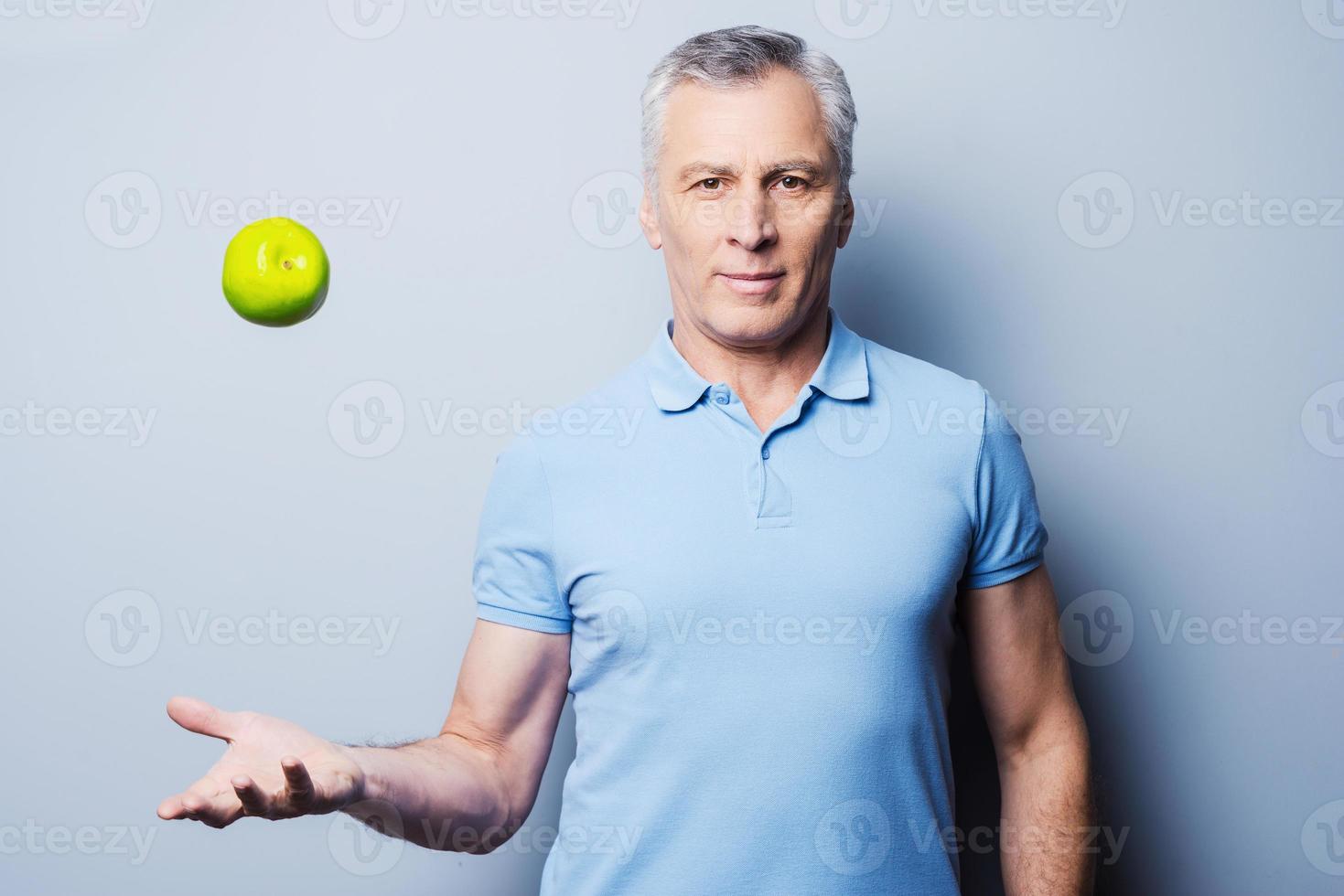 comida saudável para o sucesso. homem sênior confiante em casual jogando uma maçã verde para cima e sorrindo em pé contra um fundo cinza foto