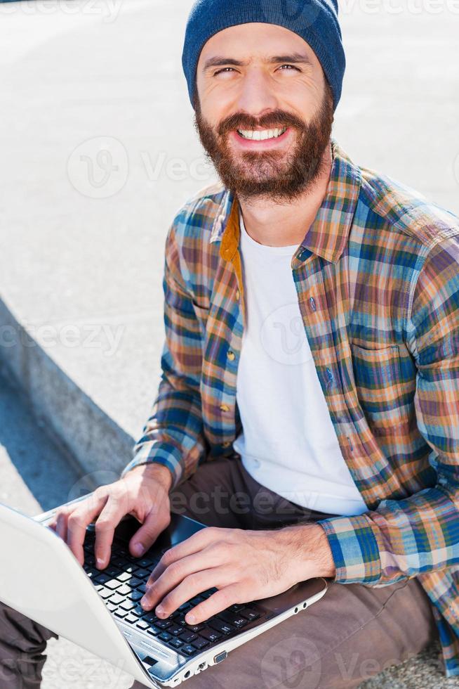 um bom dia para trabalhar ao ar livre bonito jovem barbudo sorrindo enquanto trabalhava no laptop e olhando para a câmera foto