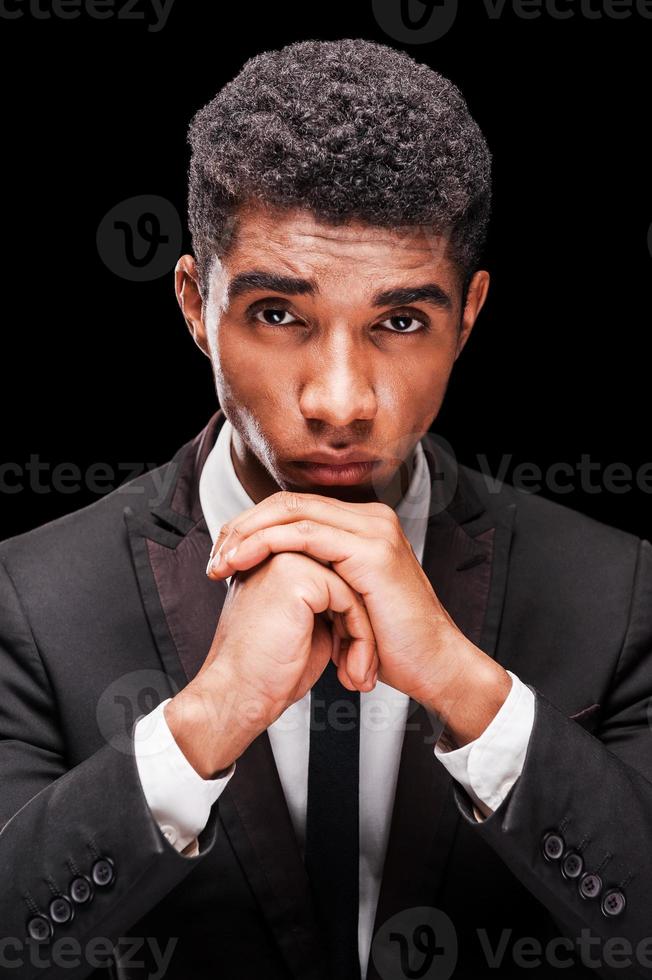 recebendo tudo. homem afro-americano sério segurando as mãos sob o queixo em pé contra um fundo preto foto