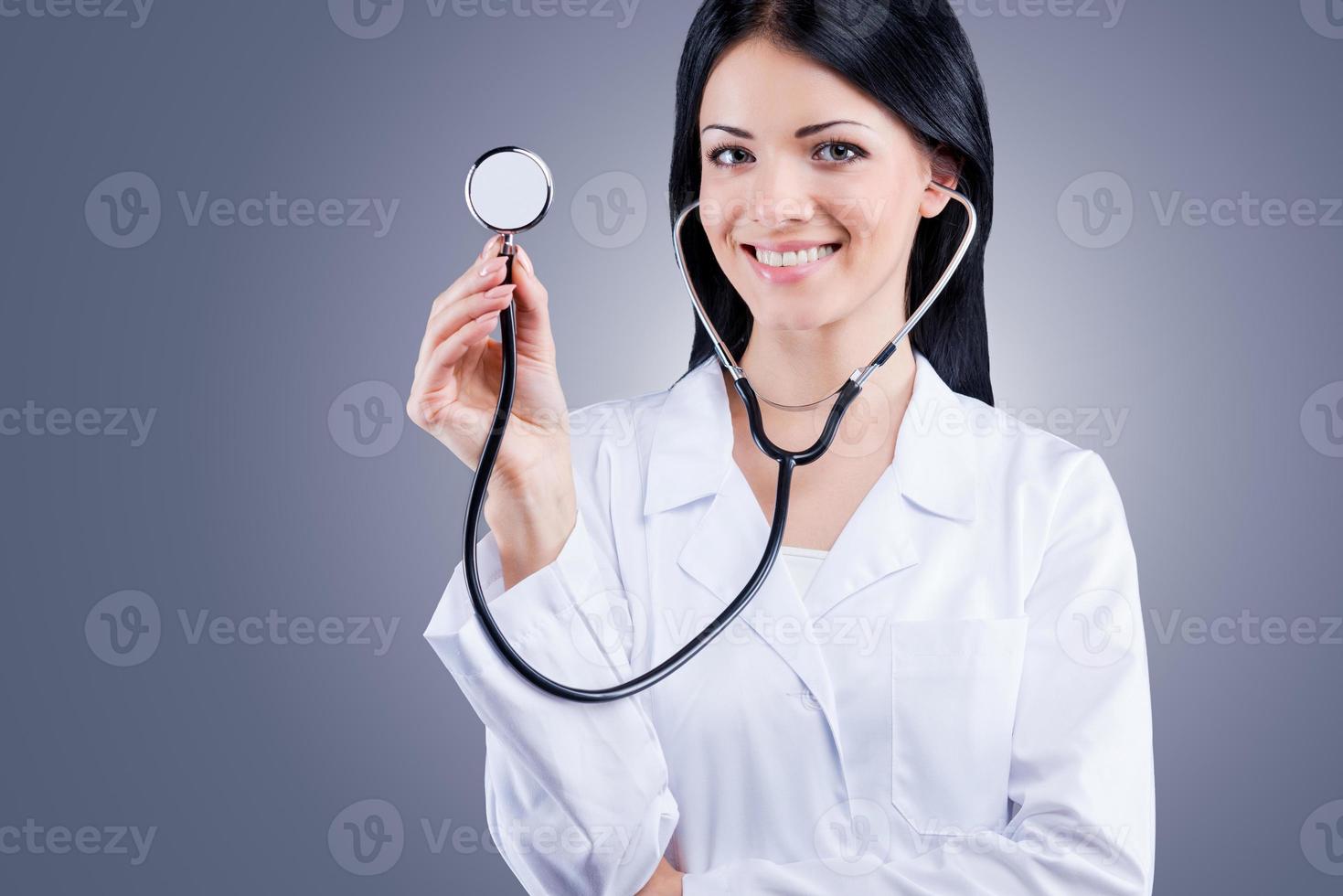 respire, por favor, médica confiante em uniforme branco segurando estetoscópio e sorrindo em pé contra um fundo cinza foto