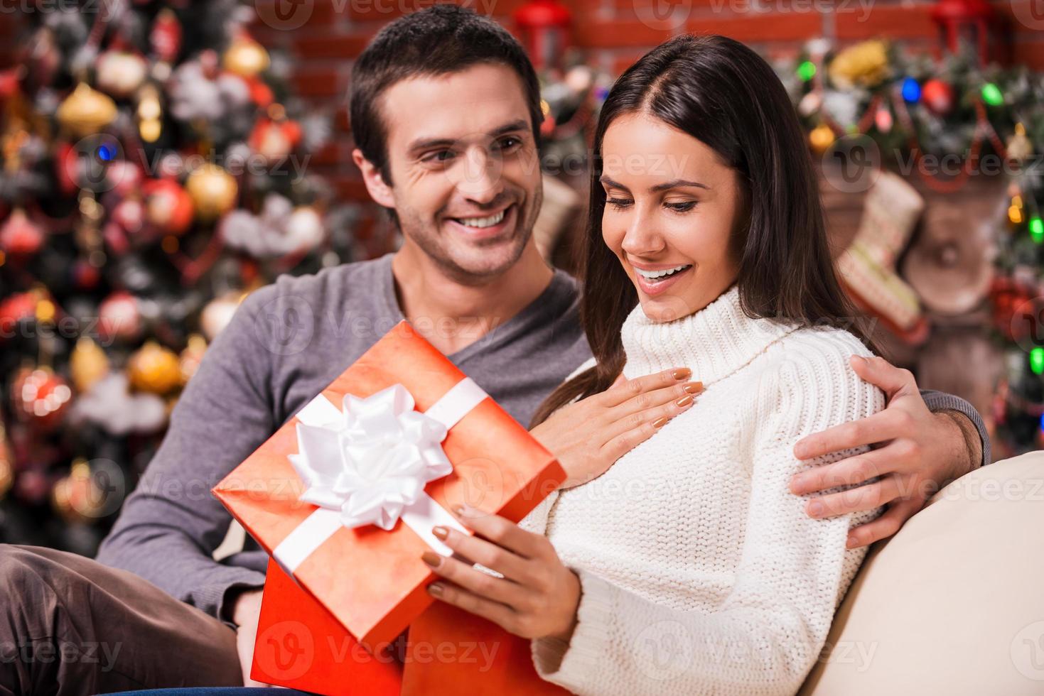 que grande surpresa bela jovem abrindo uma caixa de presente e sorrindo enquanto o namorado sentado perto dela no sofá com decoração de natal ao fundo foto