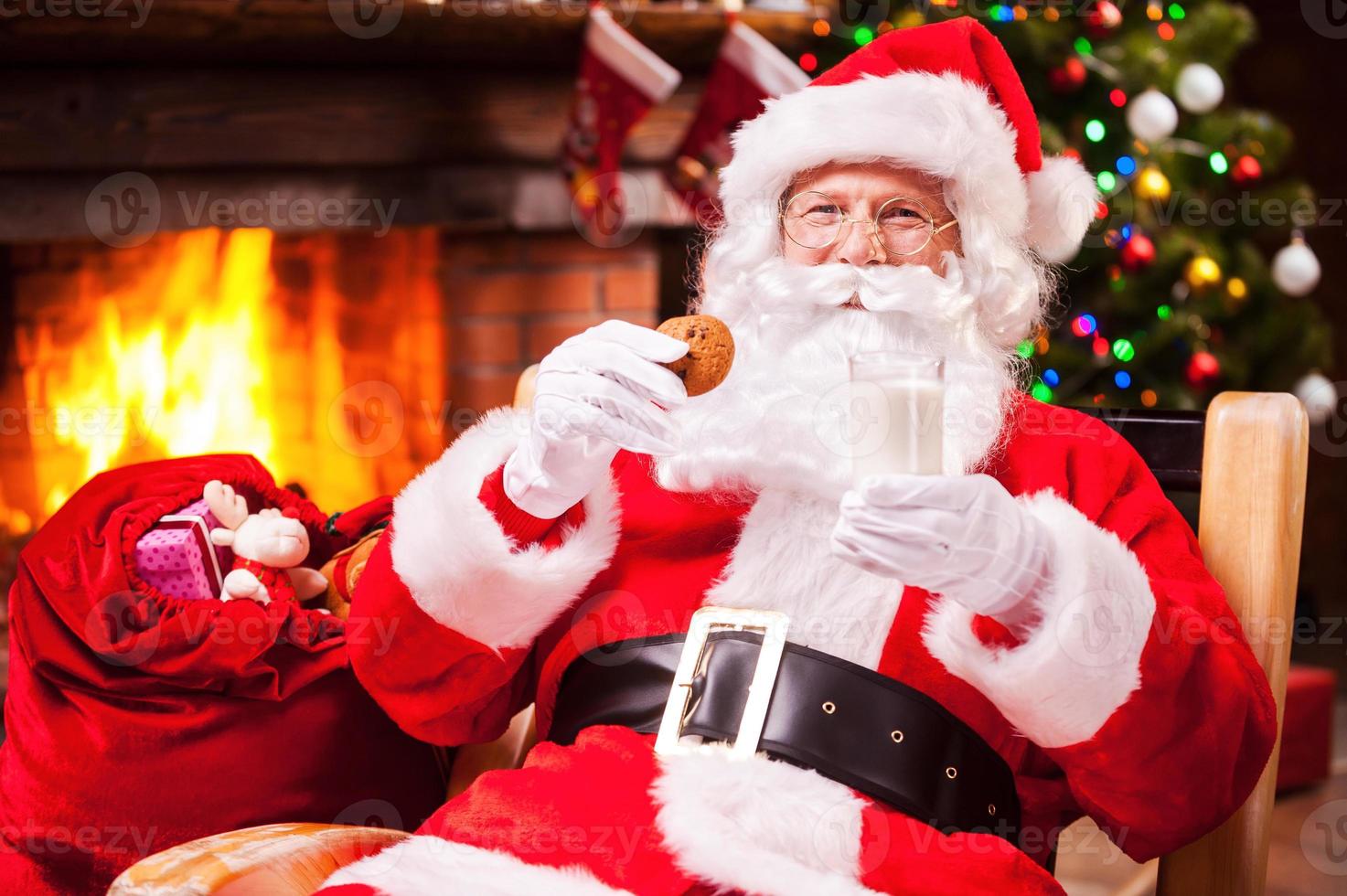 minha refeição favorita alegre papai noel segurando copo com leite e biscoito enquanto está sentado em sua cadeira com lareira e árvore de natal ao fundo foto