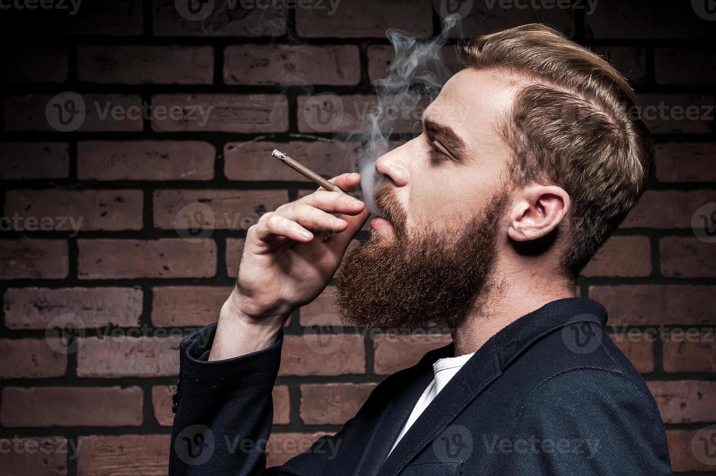 em seu próprio estilo. Vista lateral do jovem barbudo bonito fumando um cigarro em pé contra a parede de tijolos foto