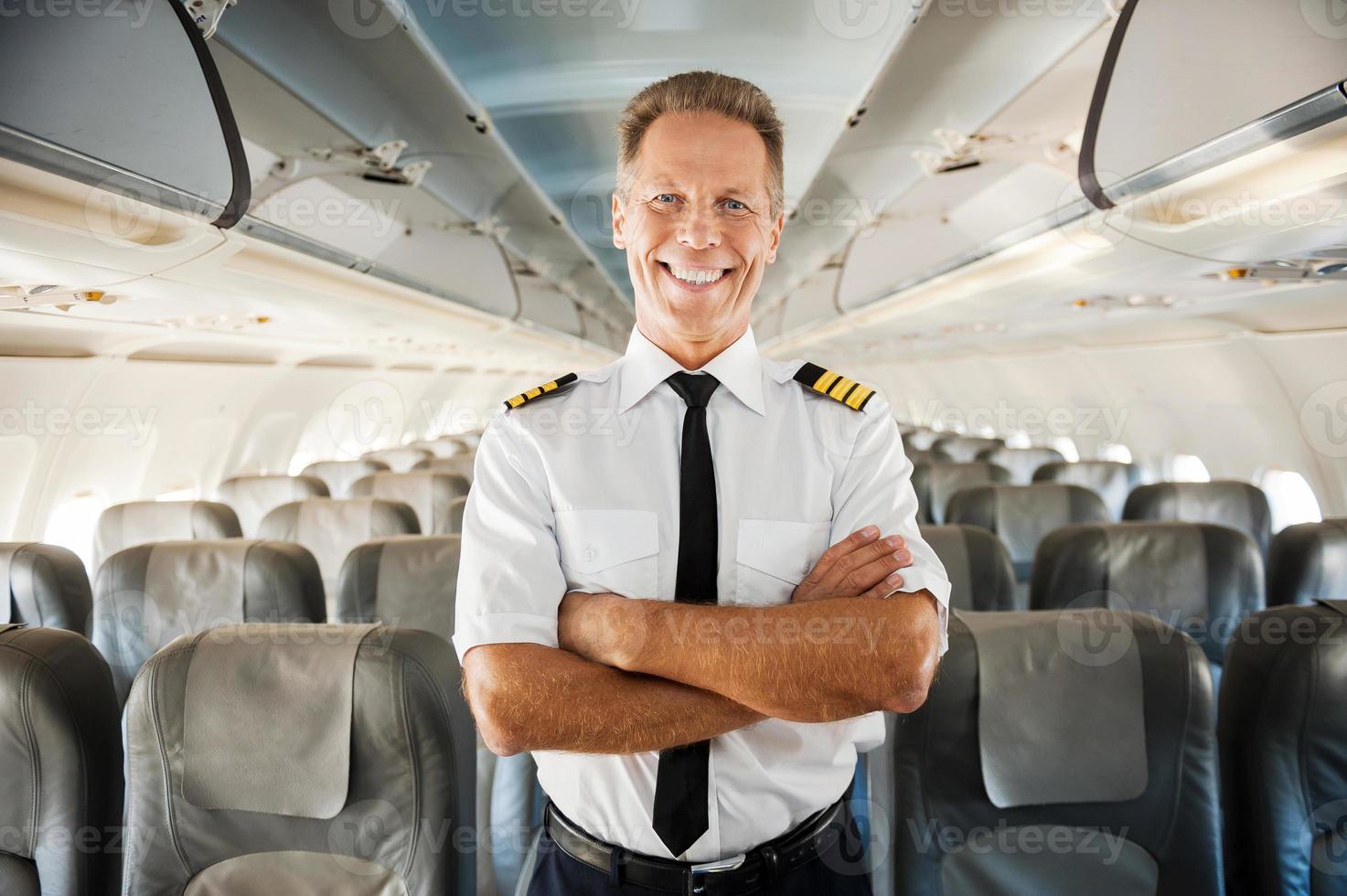 este é o meu avião. piloto masculino confiante de uniforme, mantendo os braços cruzados e sorrindo em pé dentro do avião foto