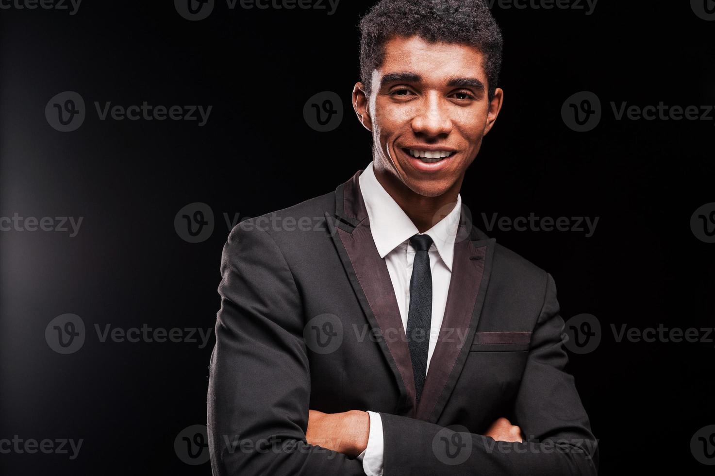 vestido de confiança. sorrindo jovem afro-americano, mantendo os braços cruzados em pé contra um fundo preto foto