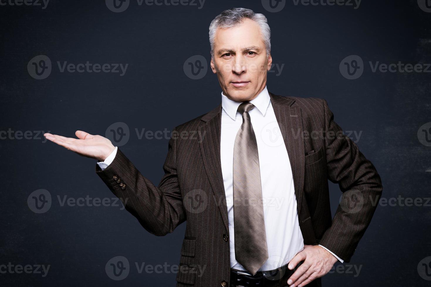 anunciando seu produto. homem sênior confiante em trajes formais, olhando para a câmera e segurando o espaço da cópia na mão em pé contra o quadro-negro foto