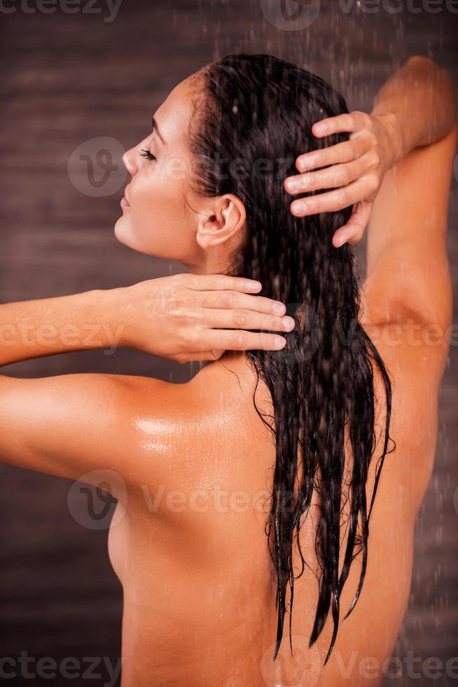 relaxante no chuveiro. vista traseira da bela jovem nua em pé no chuveiro e lavando o cabelo foto