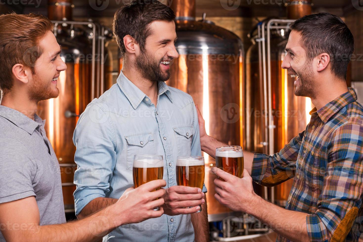 felicidades aos amigos três jovens alegres em roupas casuais conversando e sorrindo enquanto seguravam copos com cerveja e em pé na cervejaria foto