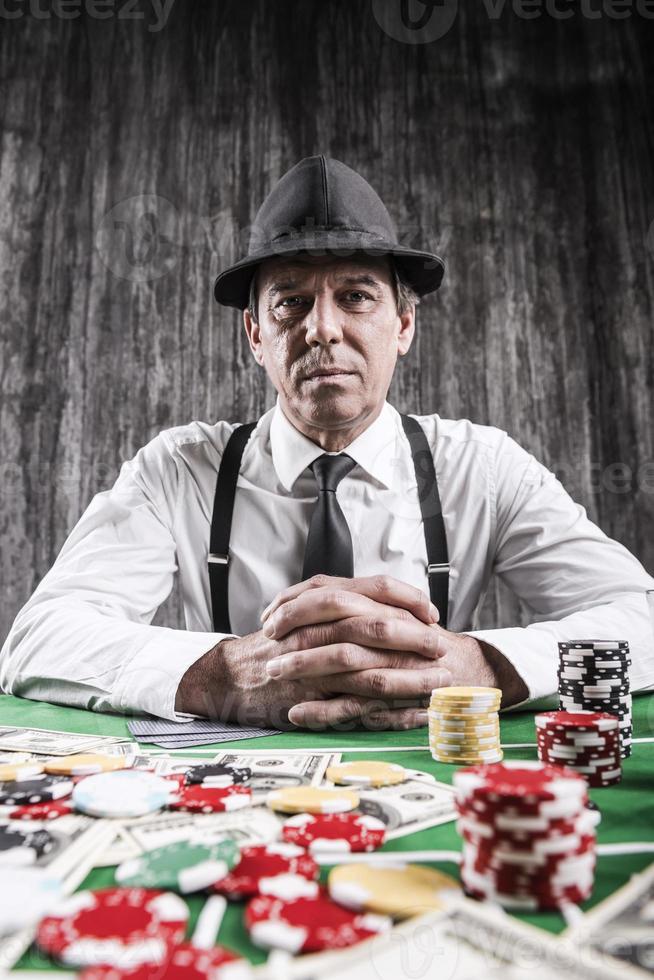 homem sênior sério na camisa e suspensórios, sentado à mesa de poker e olhando para a câmera com cartões com dinheiro e fichas de jogo ao redor dele foto
