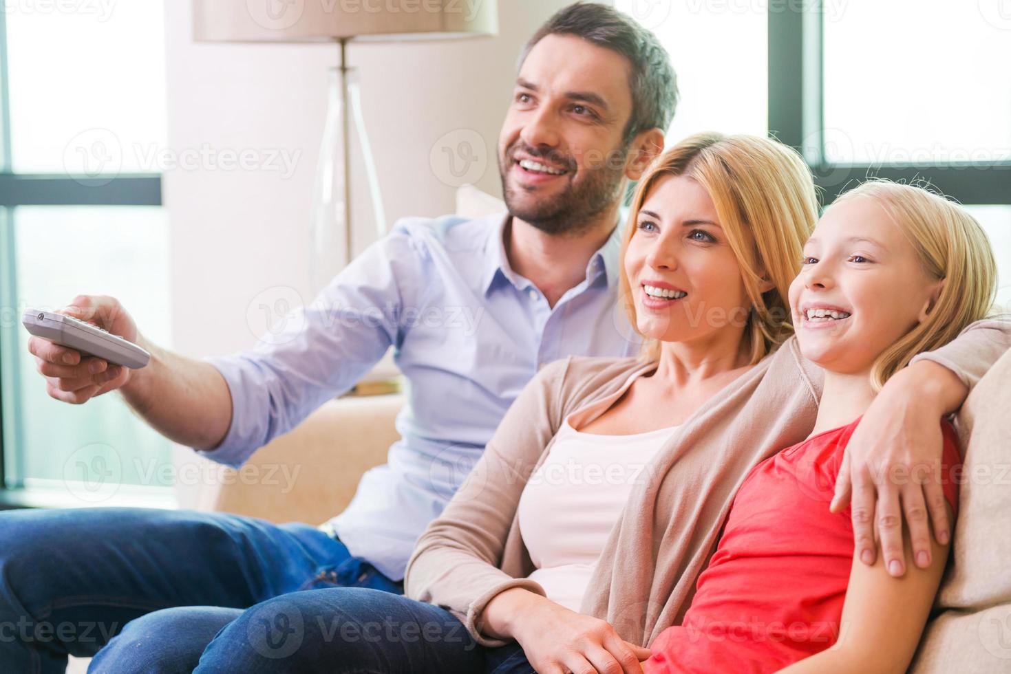 assistindo tv juntos. família feliz de três unindo-se e sorrindo enquanto está sentado no sofá e assistindo tv juntos foto