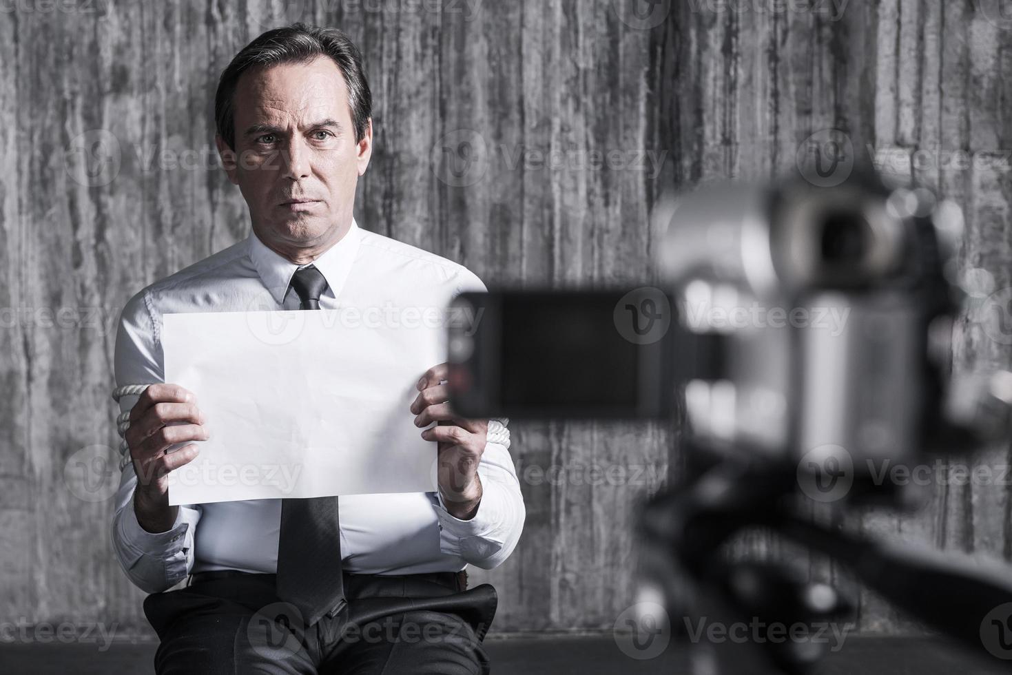 refém sem esperança. empresário frustrado pego por um criminoso sentado em frente a uma parede suja e segurando papel enquanto câmera de vídeo filmando em primeiro plano foto