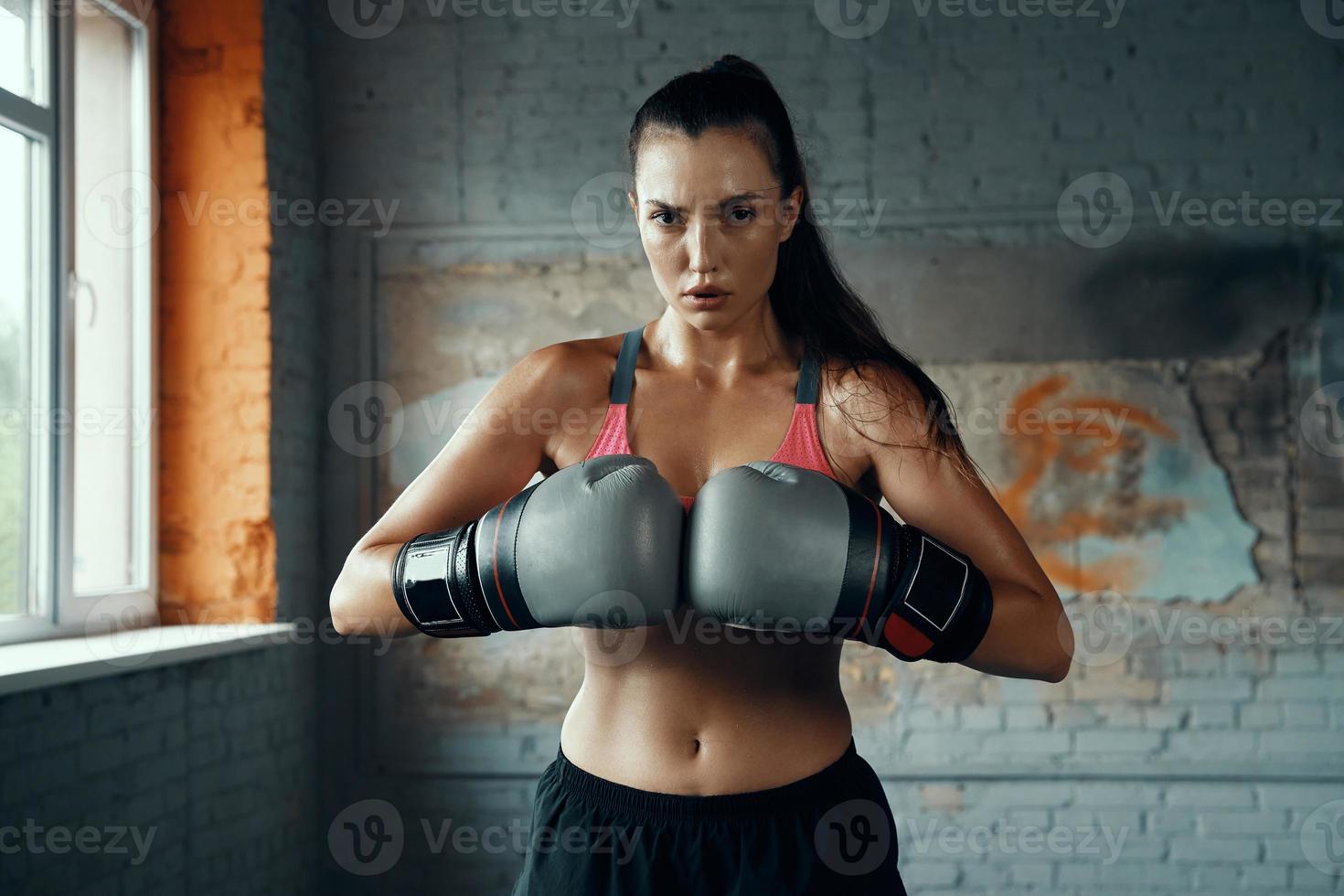 jovem confiante em luvas de boxe, olhando para a câmera em pé no ginásio foto