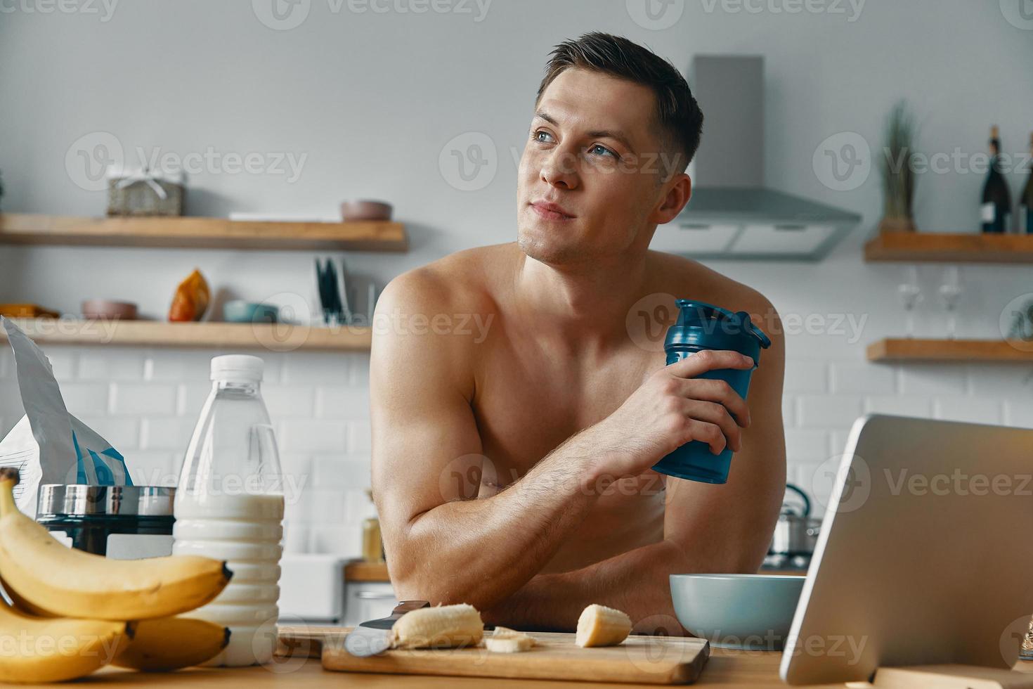 jovem pensativo desfrutando de bebida de proteína em pé na cozinha doméstica foto