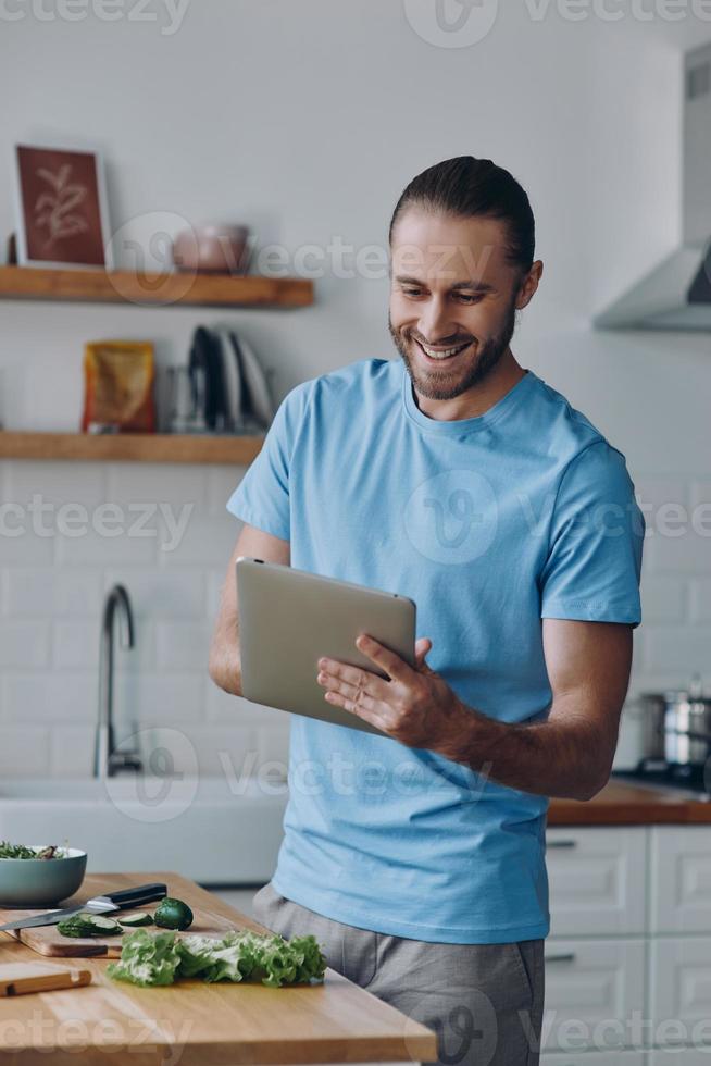 jovem alegre usando tablet digital enquanto prepara comida na cozinha doméstica foto