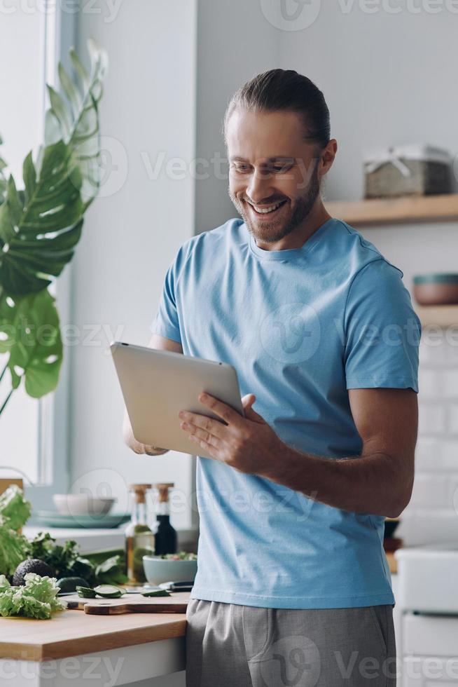 jovem feliz usando tablet digital enquanto prepara comida na cozinha doméstica foto