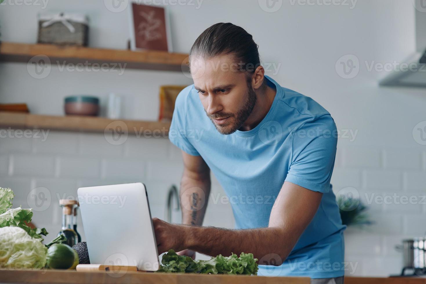 jovem bonito usando tablet digital enquanto prepara comida na cozinha doméstica foto
