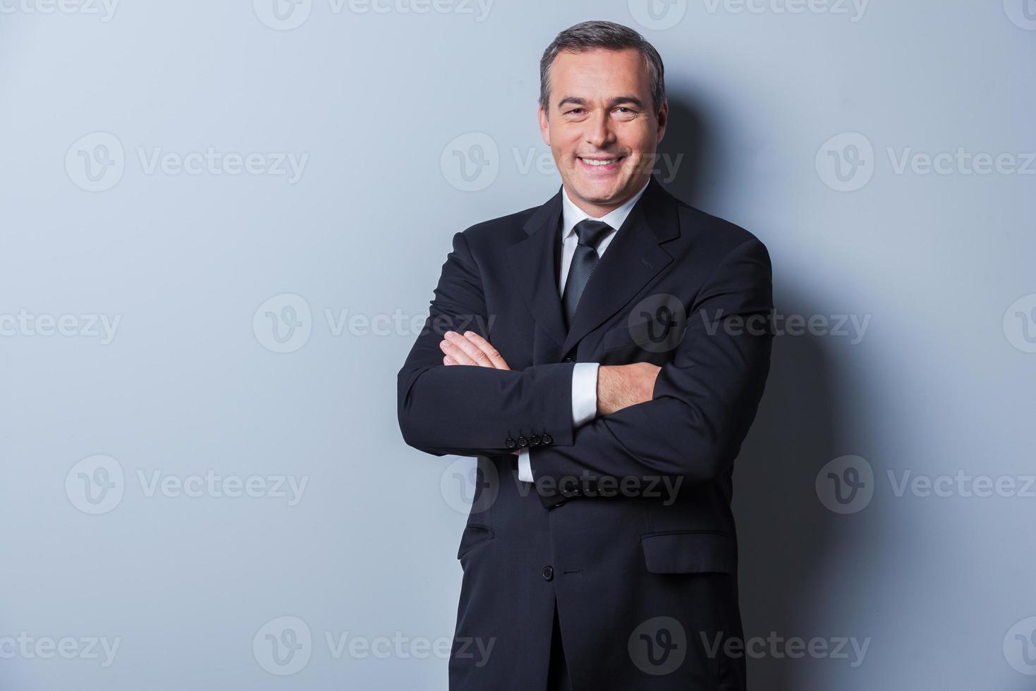 empresário de sucesso. retrato de homem maduro confiante em traje formal, olhando para a câmera e sorrindo, mantendo os braços cruzados e em pé contra um fundo cinza foto