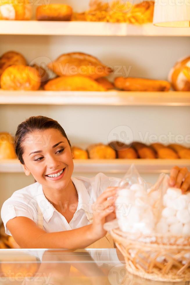 padeiro feliz no trabalho. mulher jovem e atraente no avental embalando biscoitos e sorrindo em pé na padaria foto