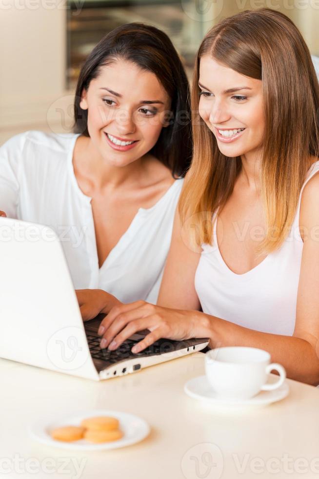 navegando na net no café. duas mulheres jovens atraentes usando computador e sorrindo enquanto estão sentados no café juntos foto