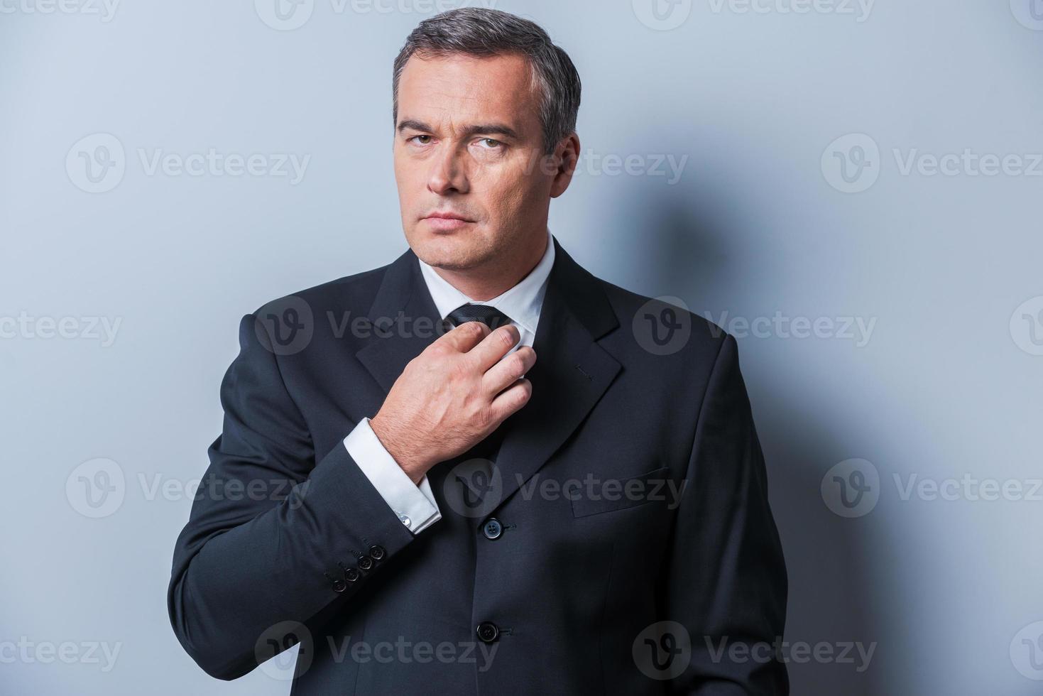 fazer o negócio parecer bom. homem maduro confiante em trajes formais, ajustando sua gravata e olhando para a câmera em pé contra um fundo cinza foto