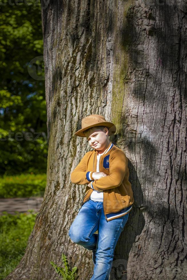 menino bonito posando com um chapéu de cowboy na floresta por uma árvore. os raios do sol envolvem o espaço. história de interação para o livro. espaço para copiar foto