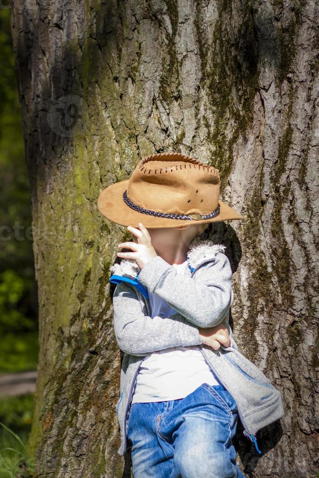 menino bonito posando com um chapéu de cowboy na floresta por uma árvore. os raios do sol envolvem o espaço. história de interação para o livro. espaço para copiar foto