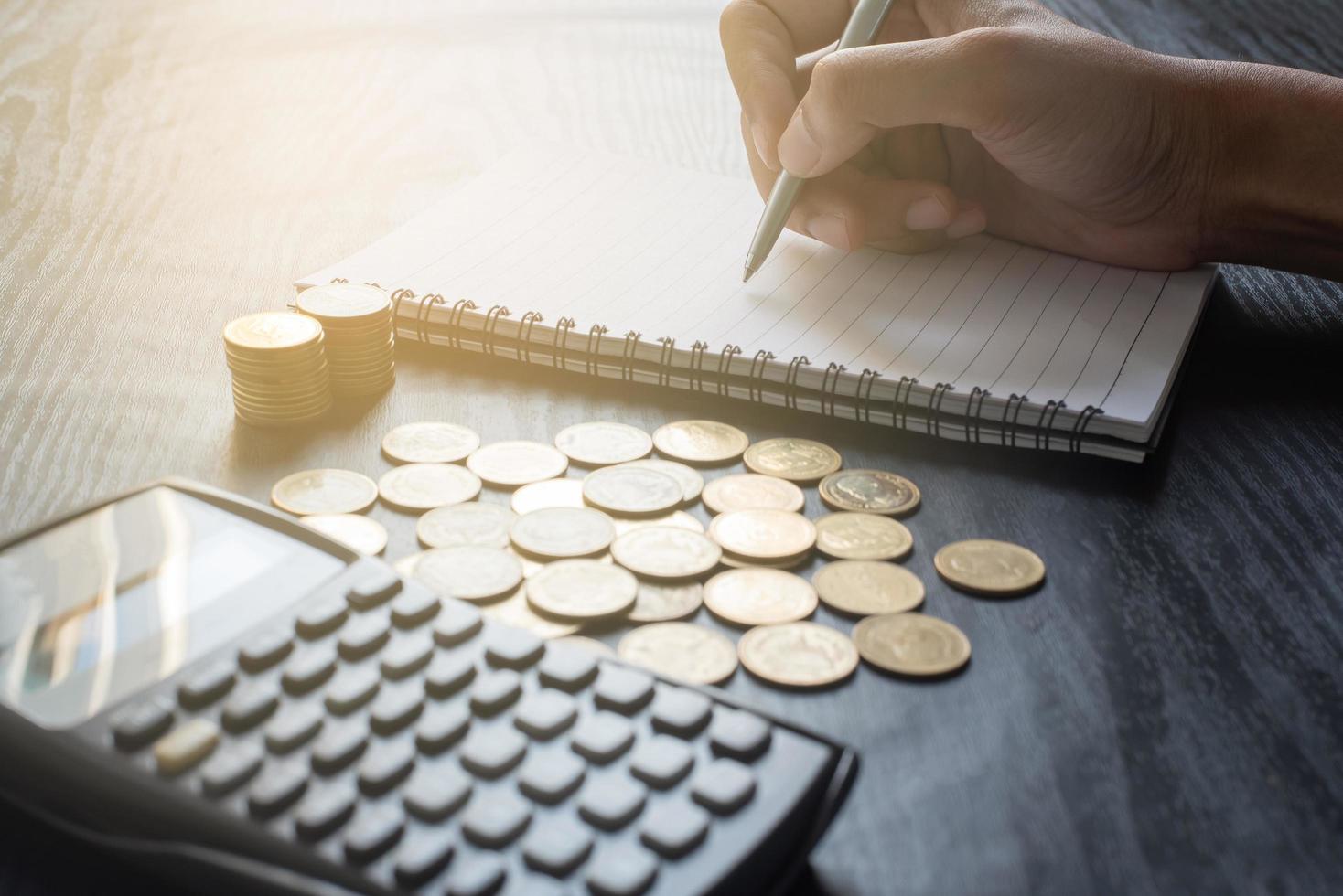 feche a mão do homem escrevendo no caderno com calculadora com moedas de ouro, caneta na mesa preta. negócios, finanças, marketing, conceito e design de comércio eletrônico foto