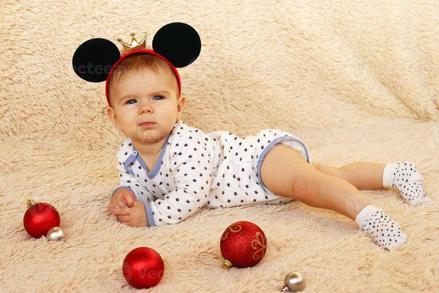 menina bonitinha com orelhas de rato em uma manta bege com bolas de natal vermelhas e prateadas. foto