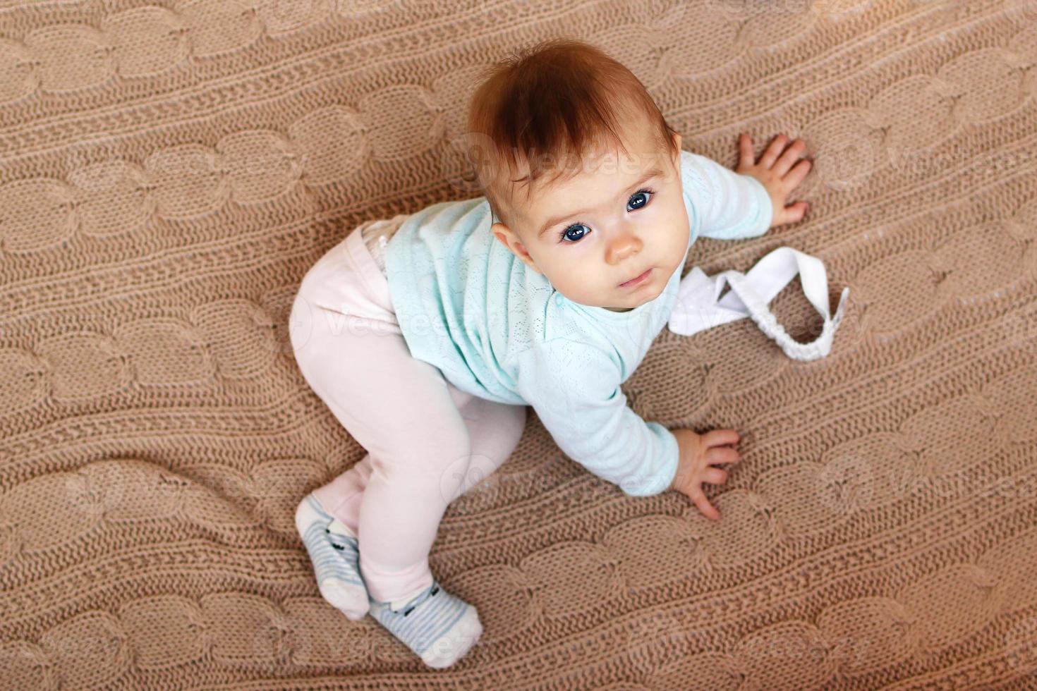 linda menina com olhos azuis e cabelo loiro está sentado em uma manta de malha bege e olhando para a câmera. foto