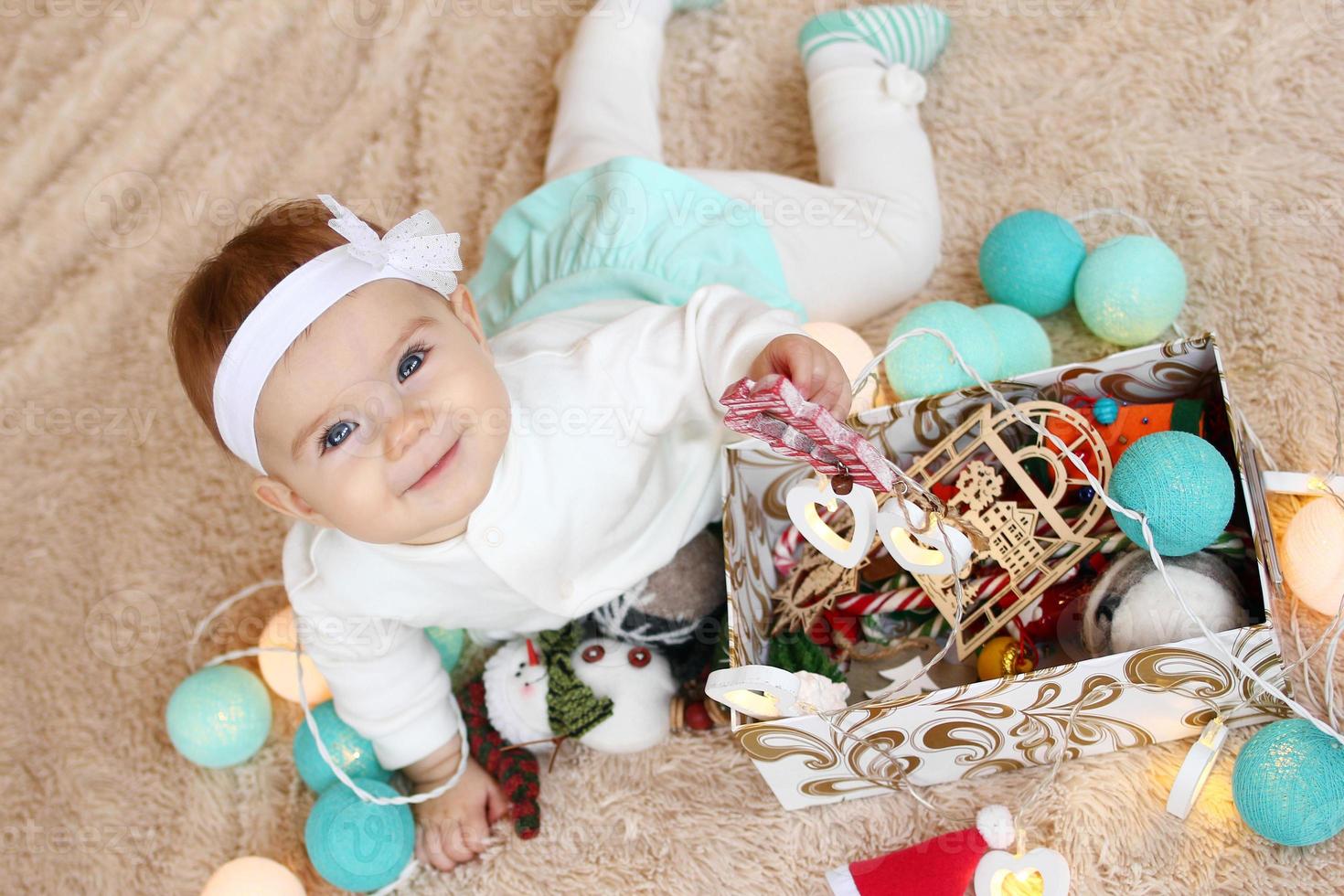 linda garotinha sorridente de vestido azul e branco está segurando o brinquedo de madeira vermelho na mão em uma manta bege com decorações de natal. foto