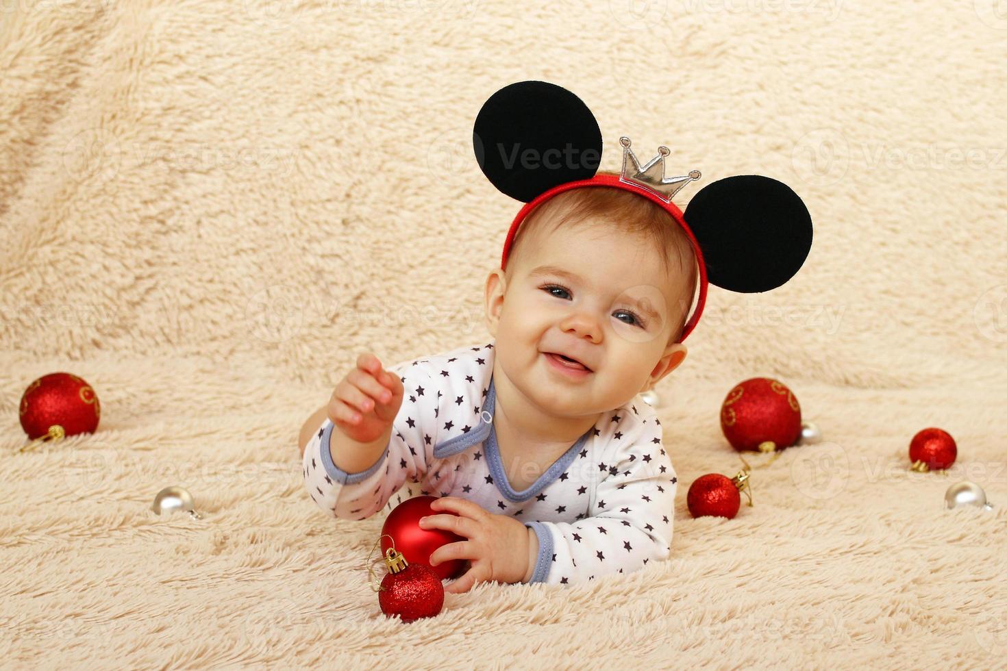 adorável menina sorridente com orelhas de rato está deitada em uma manta bege e brincando com bolas de natal vermelhas brilhantes. foto