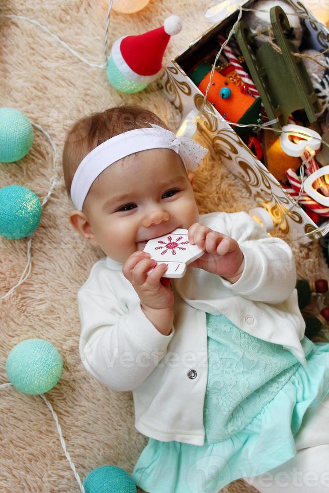 linda garotinha sorridente de vestido azul e branco está brincando com um brinquedo de madeira em uma manta bege com luzes e enfeites de natal, vista superior. foto