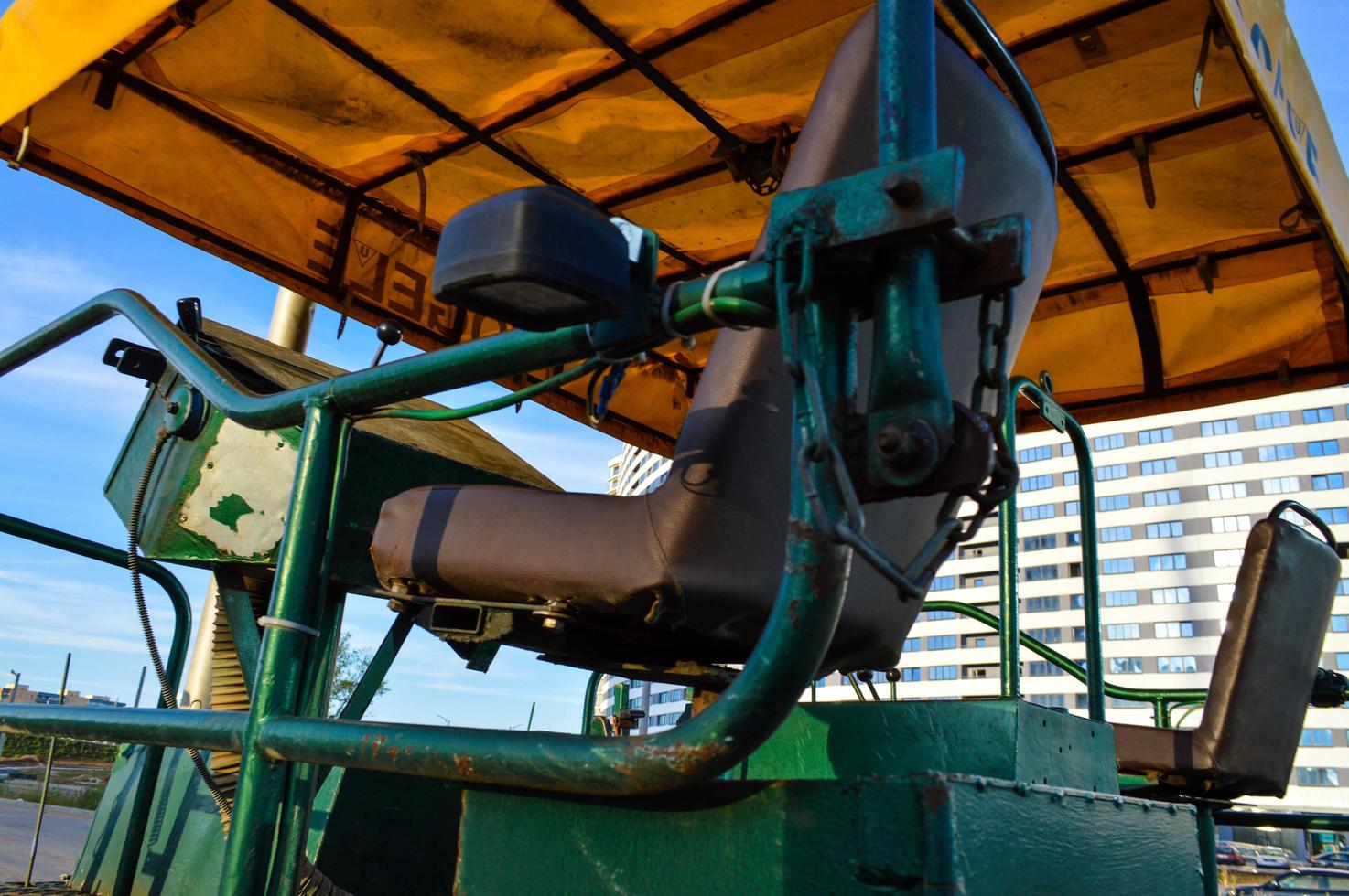 um carro para colocar asfalto fica no contexto de uma casa azul e branca. o banco do condutor é castanho, a caixa de velocidades e a estrutura metálica pintadas de verde foto