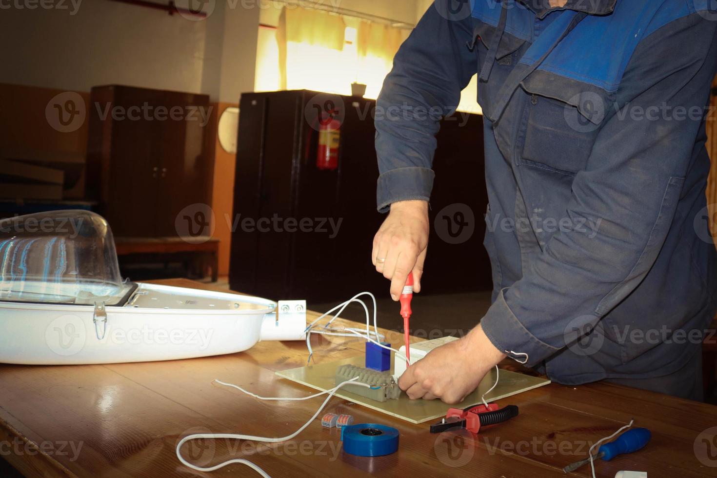 um homem que trabalha eletricista trabalha, coleta o circuito elétrico de uma grande lâmpada de rua branca com fios, um relé em uma fábrica de planta industrial foto