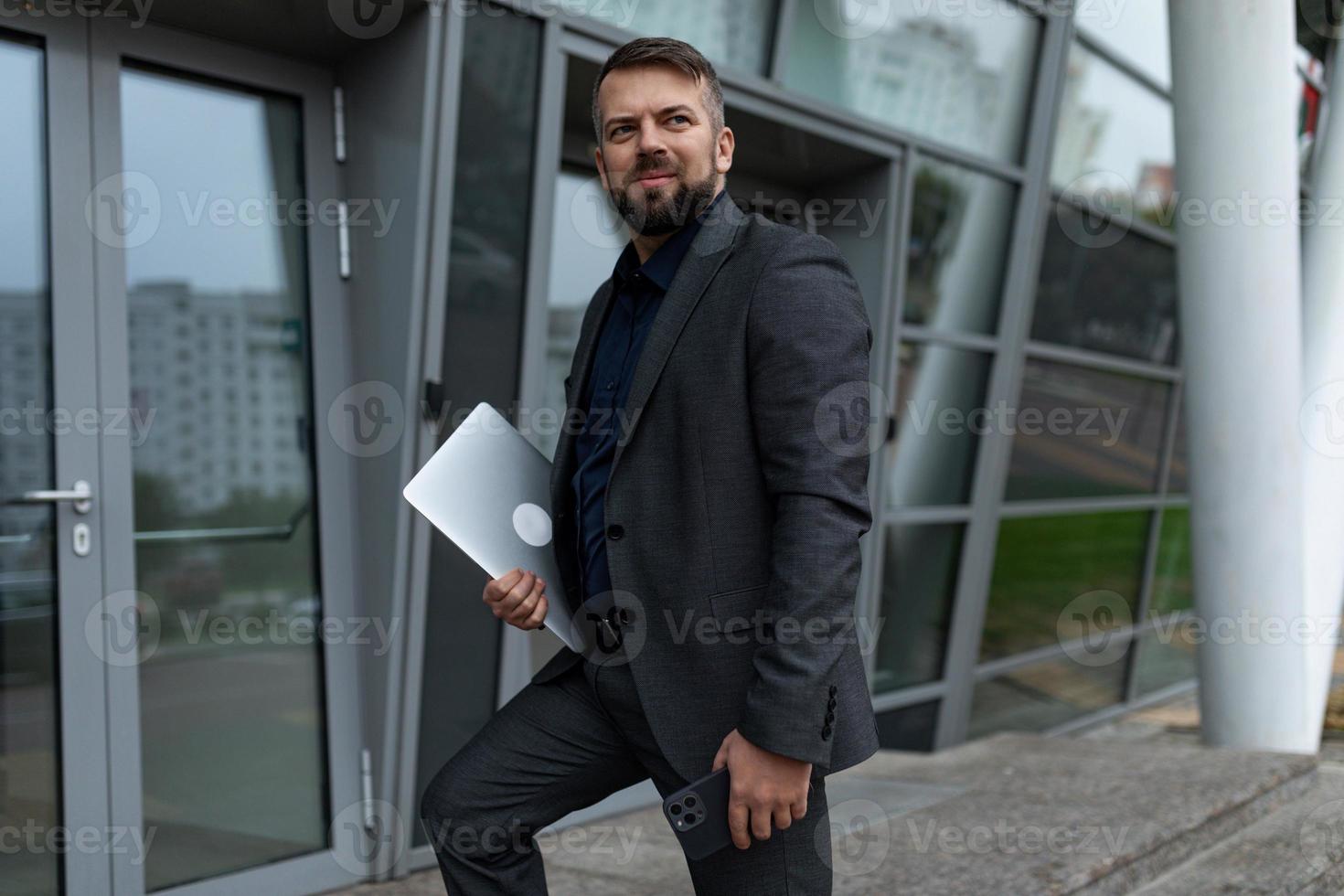 homem funcionário do banco com um laptop entra no prédio com um sorriso no rosto, conceito de sucesso nos negócios e crescimento de carreira foto