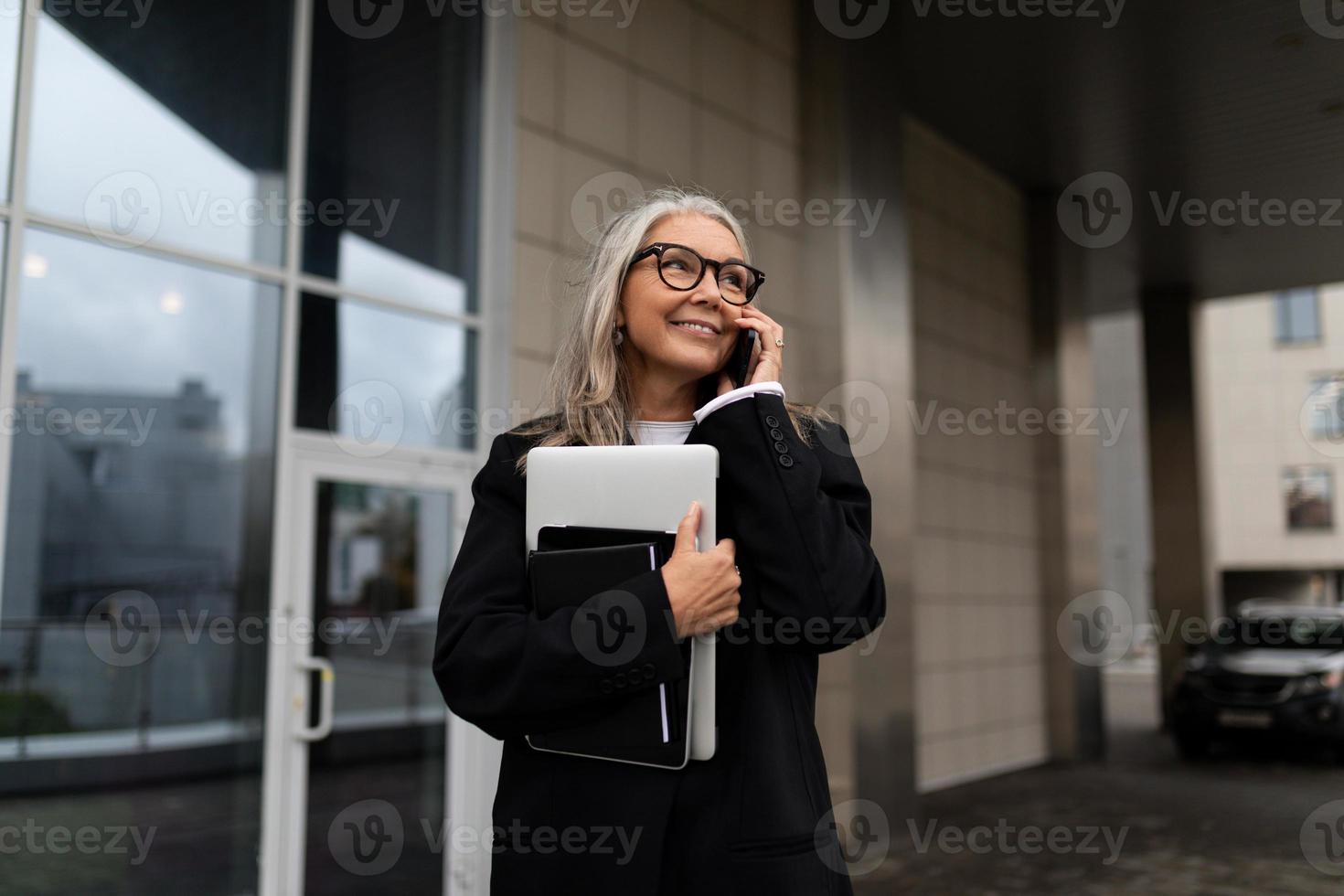 uma mulher idosa de negócios fala em um telefone celular com um laptop nas mãos contra o pano de fundo de um prédio de escritórios foto