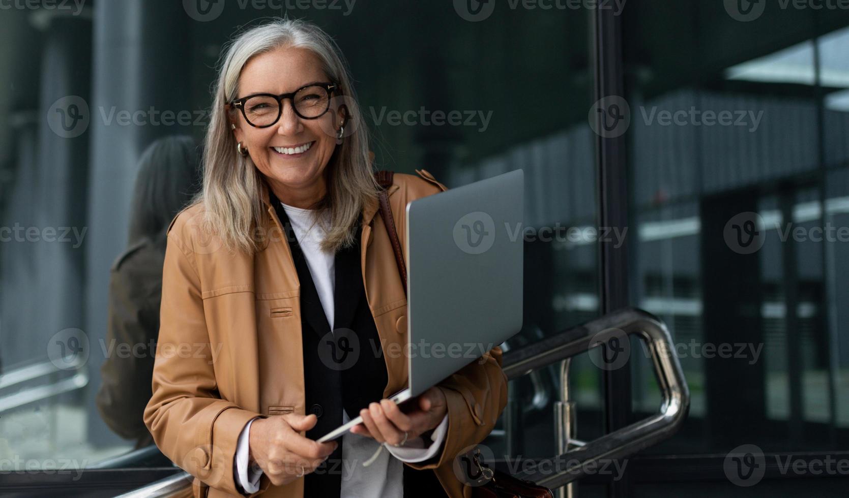 empresária idosa satisfeita com um sorriso largo e um laptop nas mãos contra o pano de fundo do centro de escritórios foto