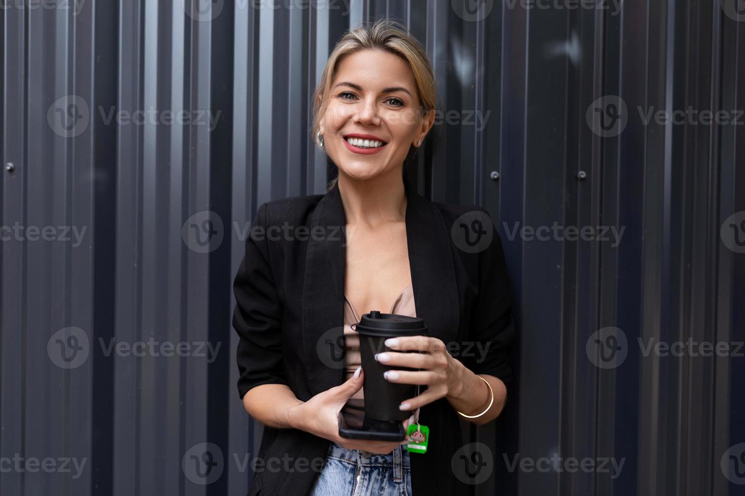 empregado de escritório de mulher bem sucedida com um telefone celular e uma xícara de chá no fundo de uma parede cinza olha para a câmera com um sorriso foto