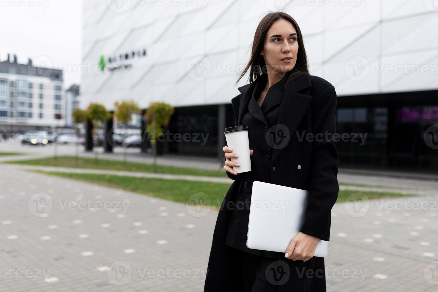 retrato de meio comprimento de uma empresária elegante com um laptop e um copo de café no contexto da fachada de um prédio de escritórios moderno foto