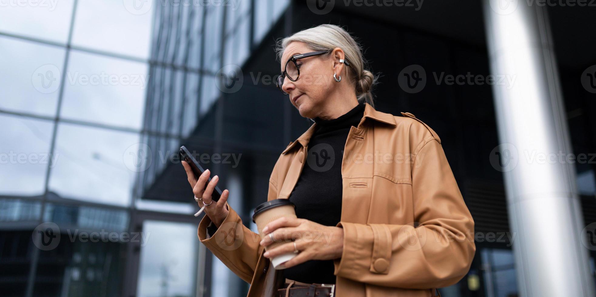 mulher idosa elegante com um telefone celular e um copo de café no fundo de um centro de negócios moderno, conceito de crescimento de carreira foto