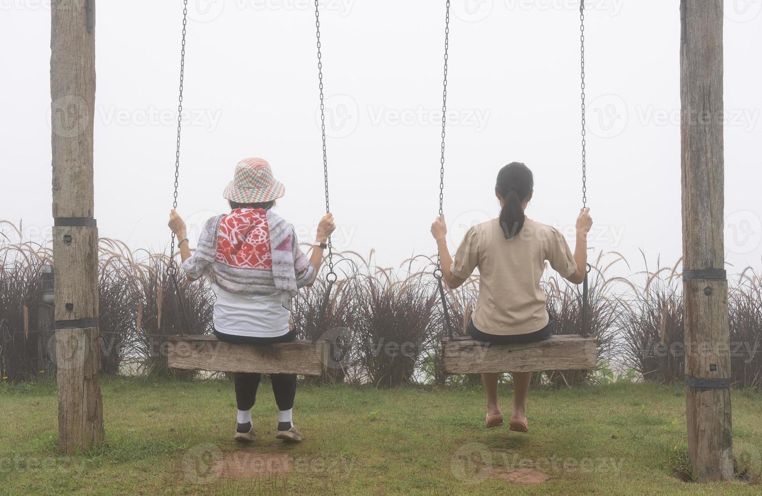 vista traseira da mulher sênior sentada e filha sentadas juntas em balanços no jardim no dia enevoado foto