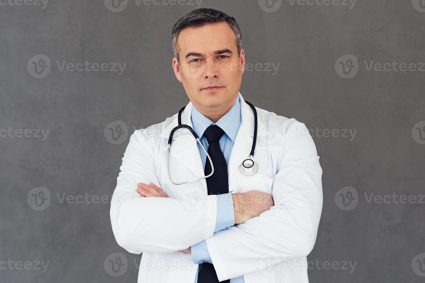 você pode confiar neste médico. médico masculino maduro, mantendo os braços cruzados e olhando para a câmera em pé contra um fundo cinza foto