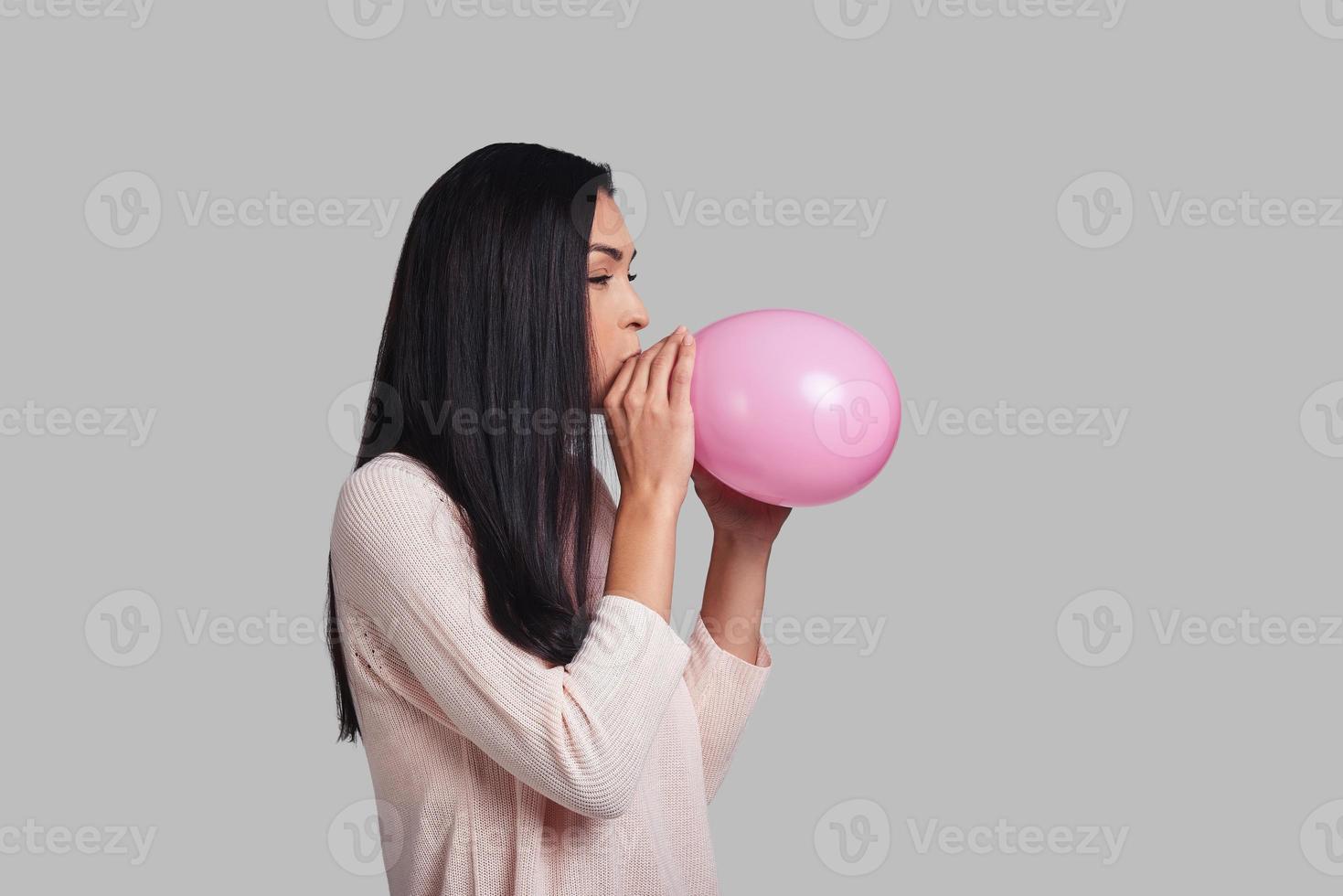 em um clima para um grande feriado. tiro de estúdio de mulher jovem e atraente em roupas casuais, explodindo um balão rosa em pé contra um fundo cinza foto