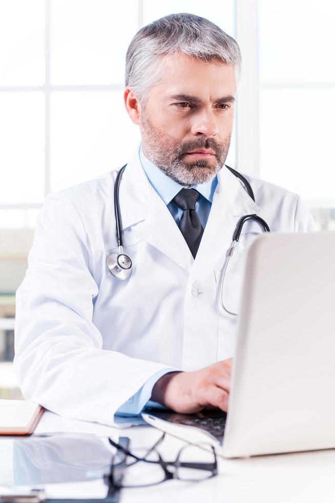clínico geral. médico de cabelos grisalhos maduros trabalhando no laptop enquanto está sentado em seu local de trabalho foto