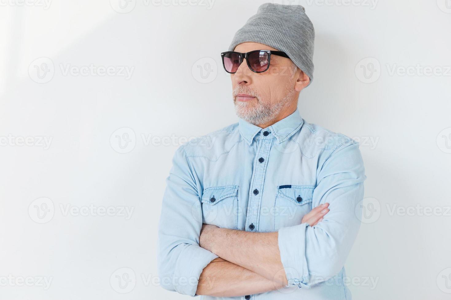 confiante em seu estilo. homem sênior de cabelos grisalhos confiante em óculos de sol e chapéu funky olhando para longe e mantendo os braços cruzados em pé contra um fundo branco foto