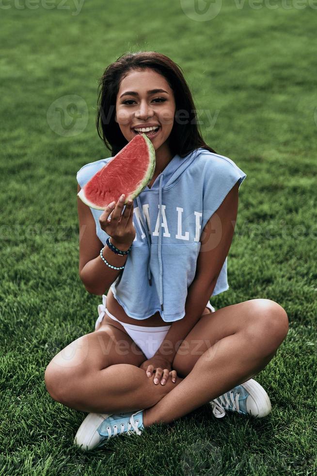 desfrutando de lanche saudável. mulher jovem e atraente comendo uma fatia de melancia e sorrindo enquanto está sentado na grama foto
