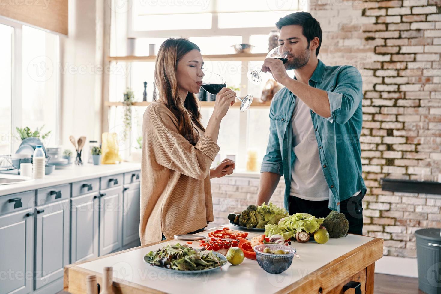 bom vinho em boa companhia. lindo casal jovem cozinhando o jantar e bebendo vinho em pé na cozinha em casa foto